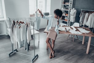 Full length of attractive young African woman talking on smart phone and holding a clothes rack while leaning on the desk in workshop