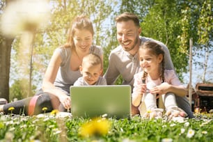 노트북을 사용하는 행복한 가족, 푸른 잔디에 앉아 있다. 봄날 초원에서 즐기는 가족.