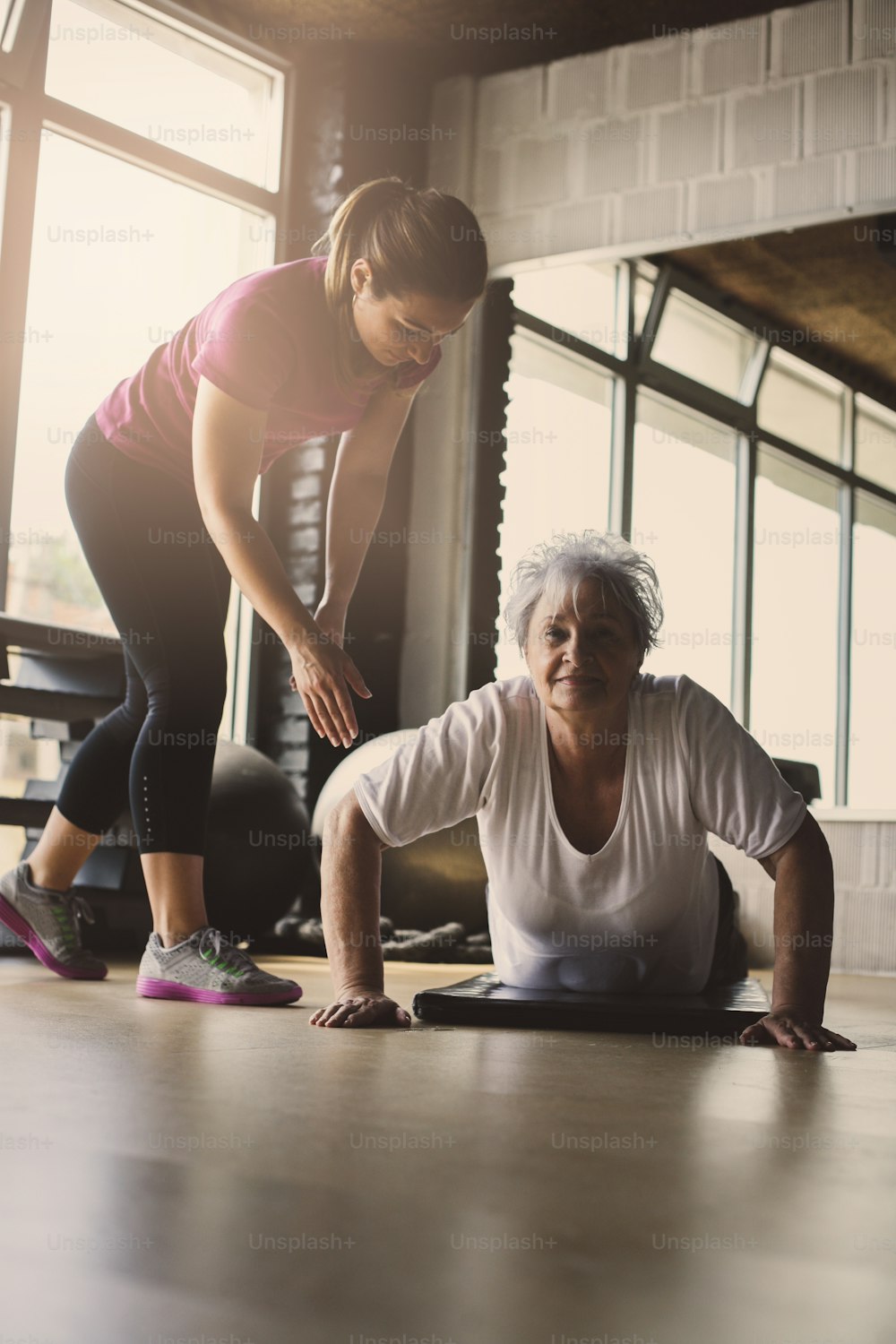 Older women doing pushups. Young personal trainer helping senior woman. Workout in rehabilitation center. Looking at camera.