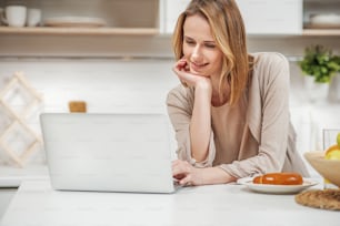 Ritratto di giovane donna gioiosa in piedi in cucina e usando il computer portatile. Lei sta sorridendo