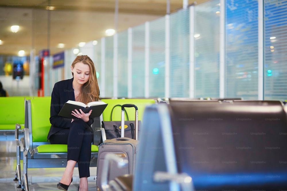 国際空港ターミナルで手荷物を持ち、フライトを待っている間に本を読んでいる若いエレガントなビジネスウーマン