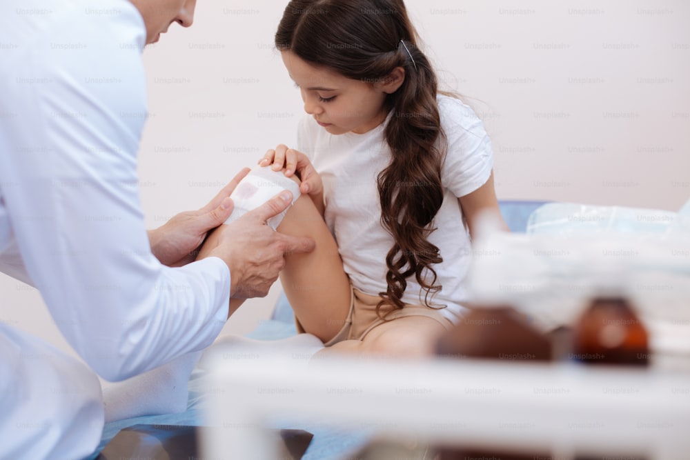 Professional help. Experienced nice male doctor sitting in front of his patient and putting a medicine dressing on her wound while giving her professional medical help