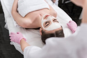 Vista superior da mulher jovem está deitada na mesa de massagem com os olhos fechados. Em seu rosto é aplicada máscara branca. Cosmetologista está de pé atrás dela. Foco seletivo