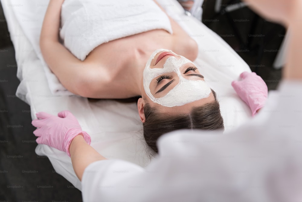 Top view of young woman is lying on massage table with closed eyes. On her face is applied white mask. Cosmetologist is standing behind her. Selective focus