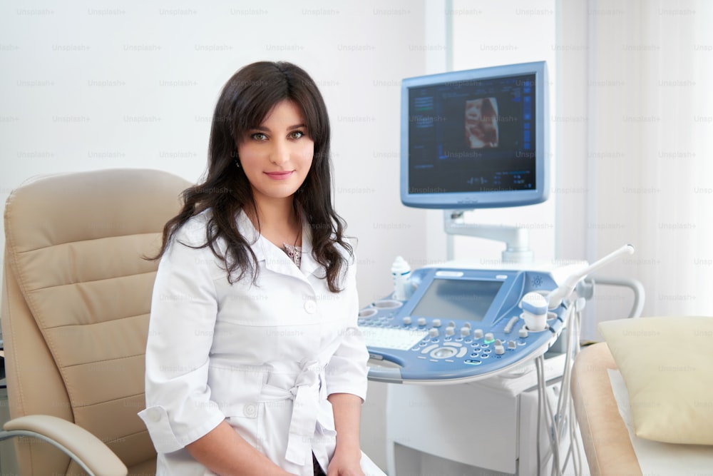 Joven y atractiva doctora, sonriendo alegremente a la cámara, sentada en su oficina cerca de la máquina de escaneo de ultrasonido, espacio de copia, ginecología, ginecología, embarazo, profesionalidad de la salud, experta.