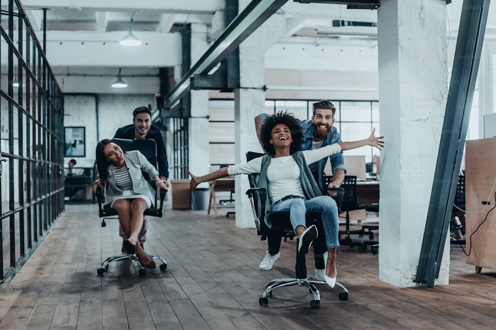 Grupo de jovens empresários alegres em roupas casuais inteligentes se divertindo enquanto correm em cadeiras de escritório e sorriem
