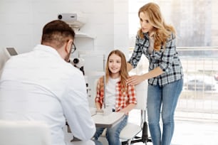 Family support. Loving mom and her pretty daughter sitting at ophthalmologists office and waiting for his verdict while he studying results of girls tests