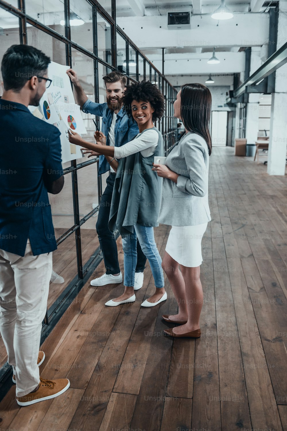 Longitud completa de jóvenes modernos en la estrategia comercial de planificación de ropa casual inteligente mientras que la mujer joven señala el papel grande que se muestra en la pared de vidrio en la oficina