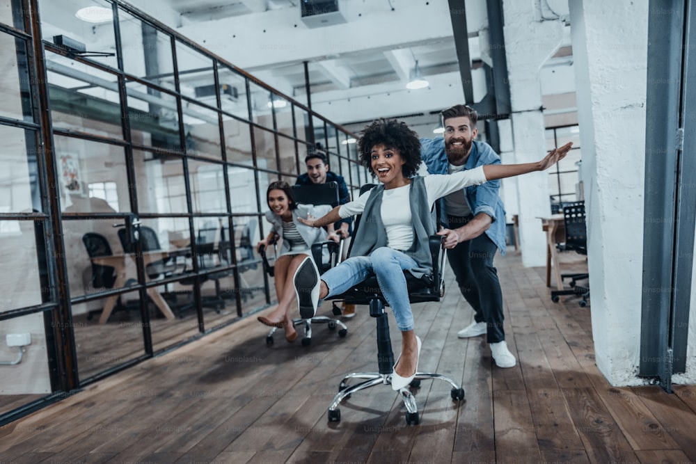 Quatro jovens empresários alegres em roupas casuais inteligentes se divertindo enquanto correm em cadeiras de escritório e sorriem