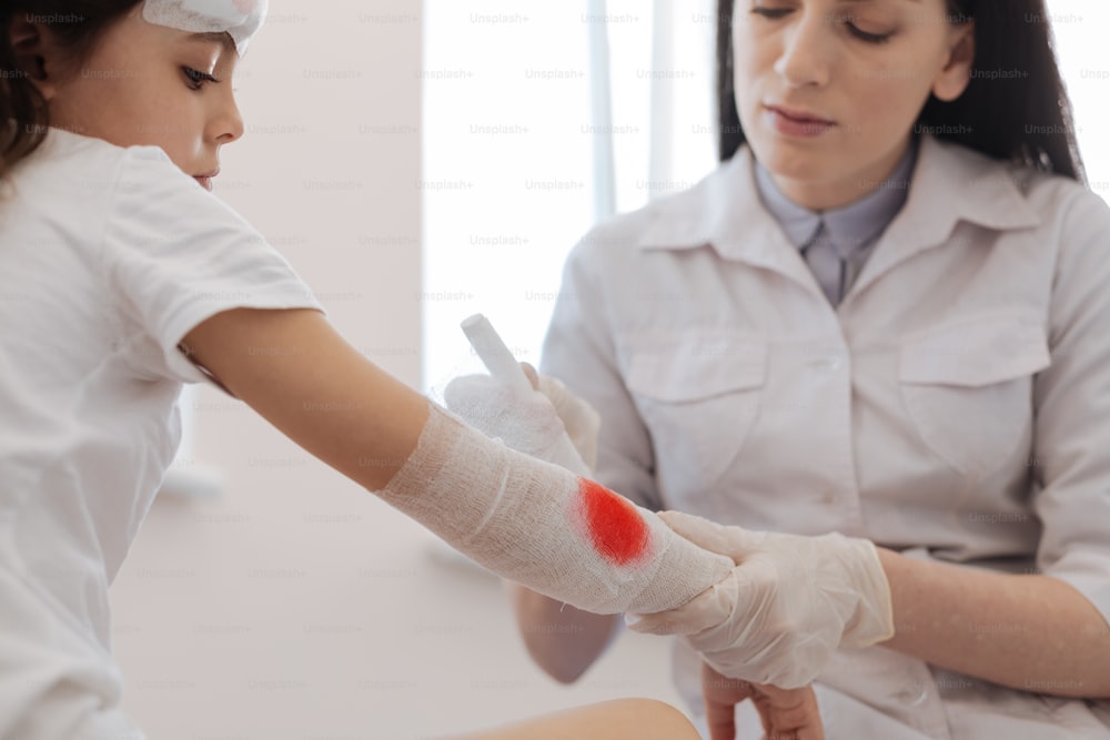 Dangerous injury. Serious nice female doctor sitting in front of her patient and wrapping the bandage around the wound while stopping bleeding