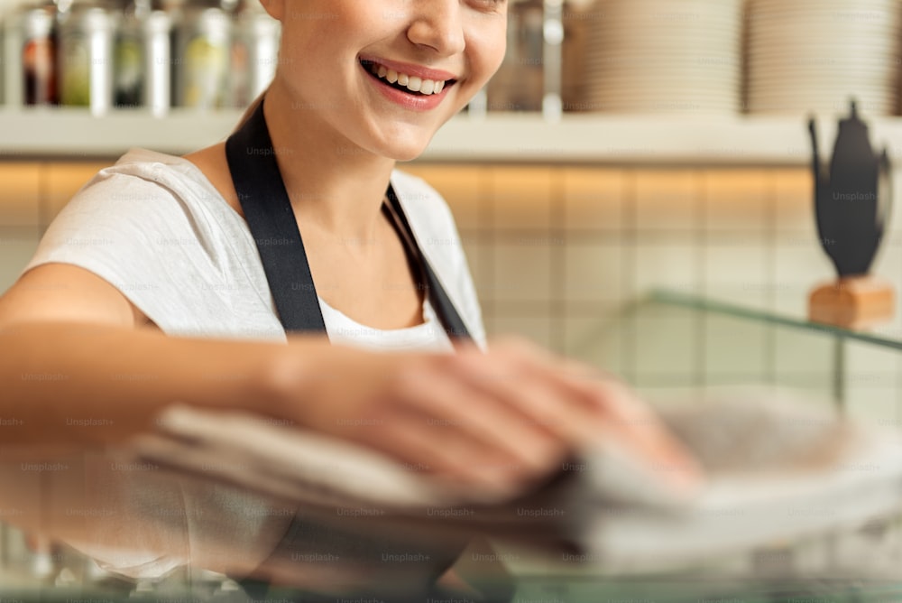 Happy waitress is cleaning glass case in cafe. She is standing and smiling