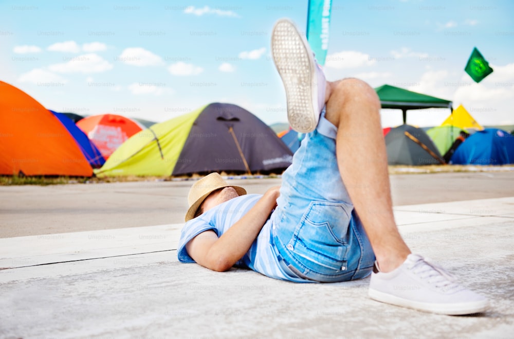 Unrecognizable man with hat on summer music festival, lying on concrete path, resting, various colorful tents behind him.