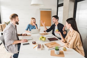 Hilarious managers are discussing project. They sitting around workplace and listening to their colleague, who is jolly gesticulating