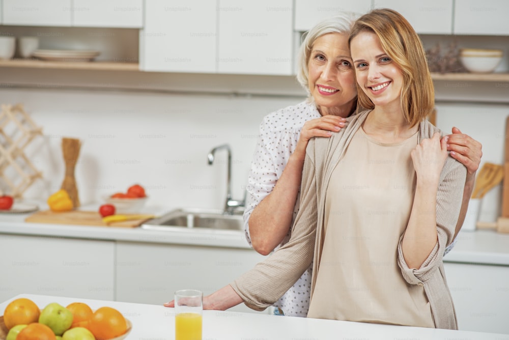 My mom is my best friend. Charming young woman and senior lady are embracing with love. They are standing in kitchen and smiling