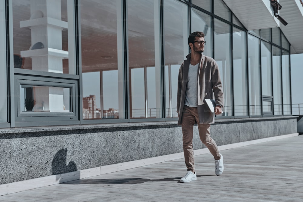 In voller Länge eines selbstbewussten jungen und gutaussehenden Mannes, der ein digitales Tablet trägt, während er im Freien spazieren geht