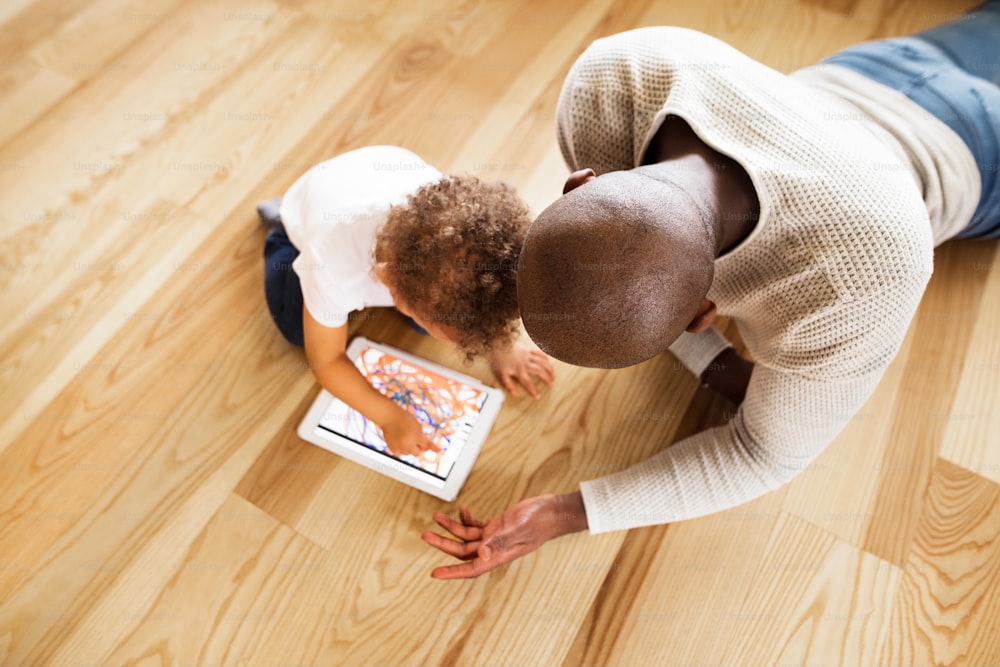 귀여운 혼혈 딸과 함께 집에 있는 젊은 아프리카계 미국인 아버지는 바닥에 누워 무언가를 보거나 태블릿으로 게임을 하고 있습니다.