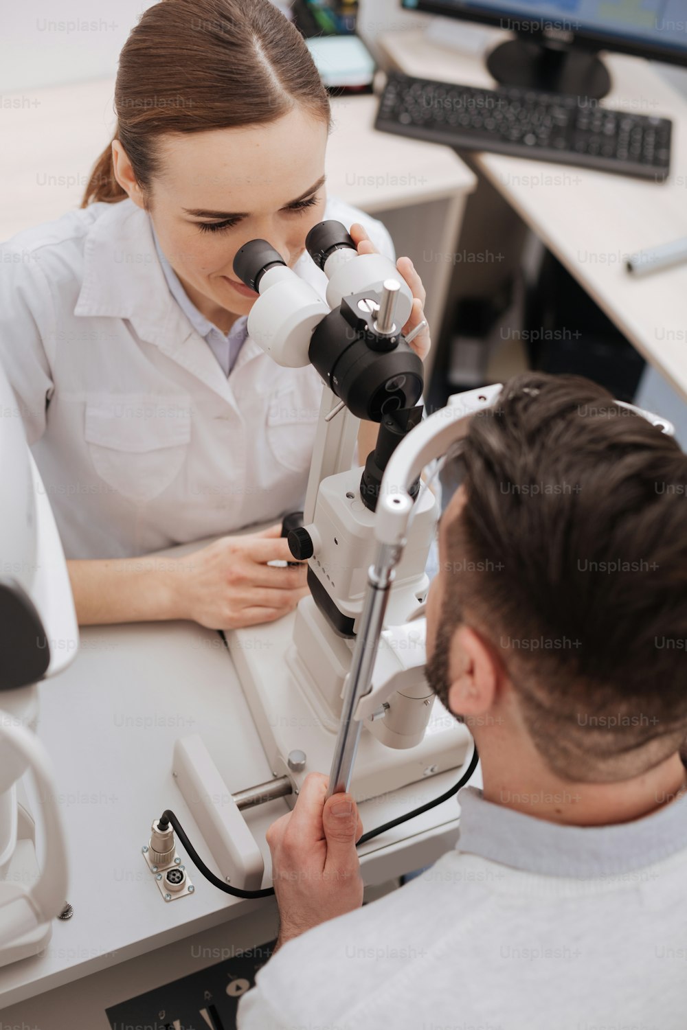 Examen oftalmológico. Vista superior de una bonita óptica profesional sentada frente a su paciente y examinando sus ojos mientras usa equipo médico