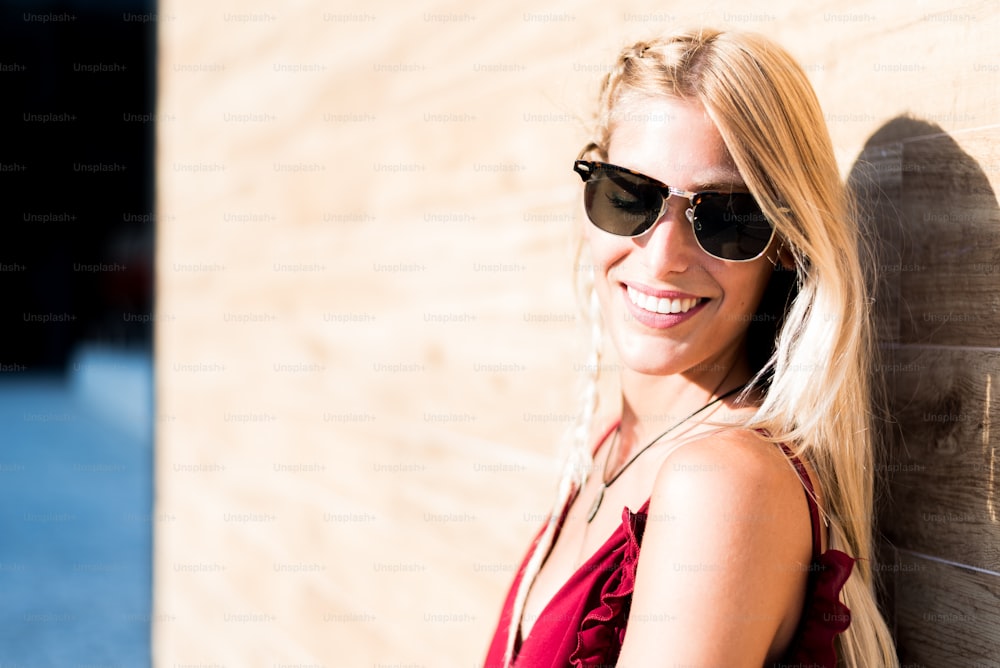 mulher loira bonita feliz que usa óculos de sol sorrindo em uma parede de madeira