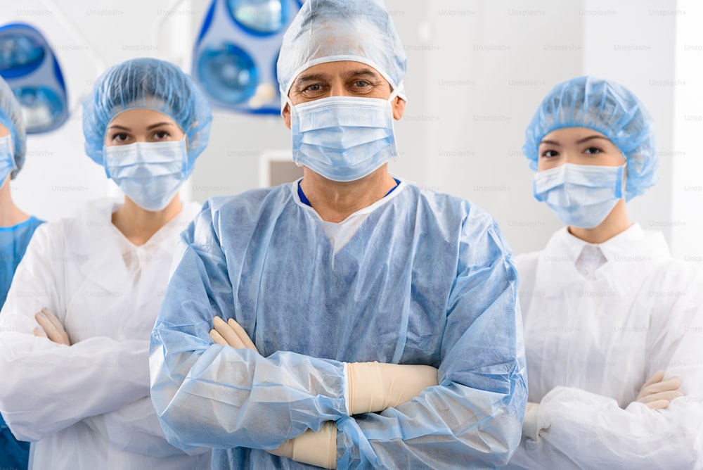 Cheerful surgeon with assistants in masks are crossing arms and looking at camera with joy