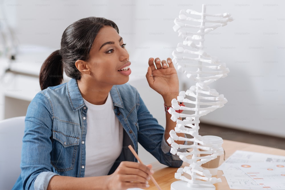 Genetic studies. Smart serious young woman sitting at the table and looking at the DNA model while studying genetics