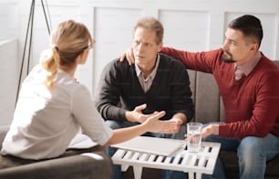 Useful advice. Unhappy cheerless sad man looking at his therapist and listening to her while sitting with his boyfriend on the sofa