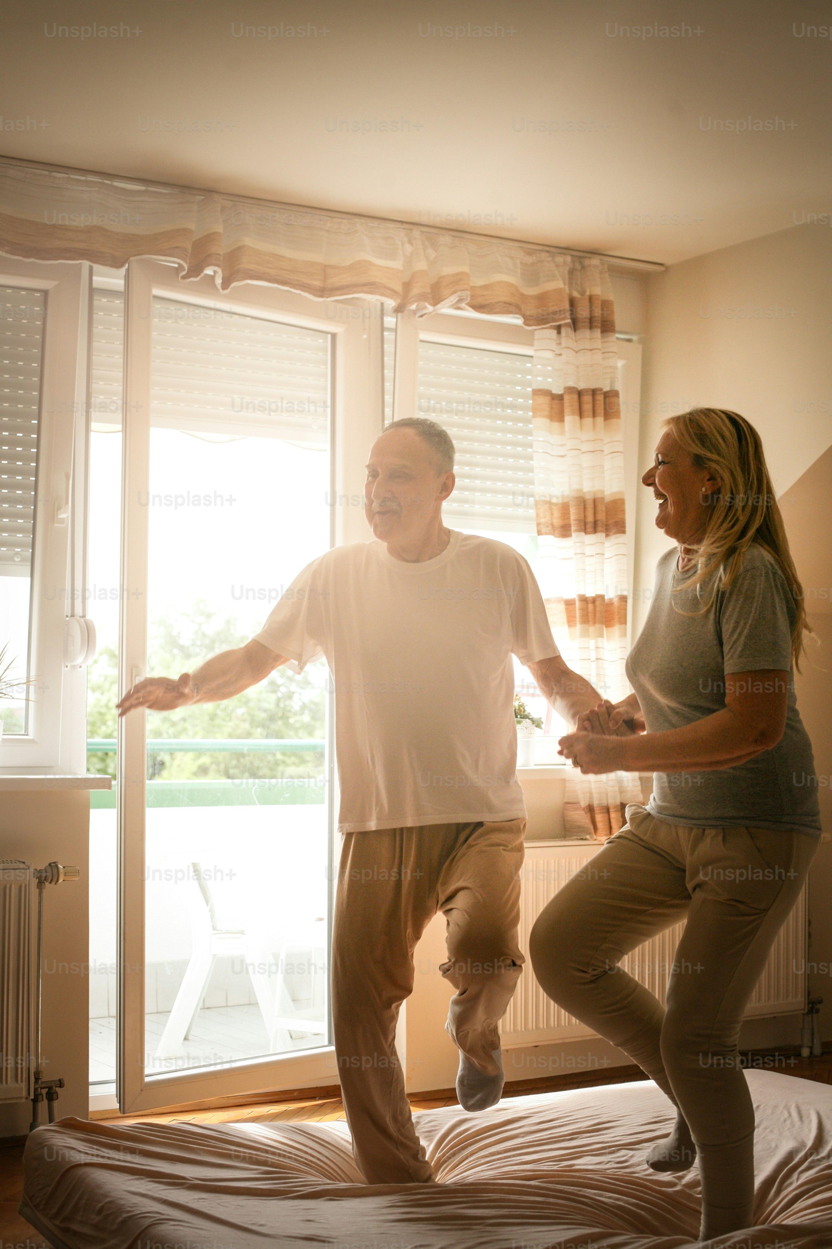 Senior couple dancing and jumping together on bed  holding hands.