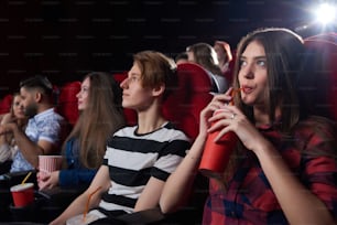 Friends and couples, watching interesting film at big cinema hall with red chairs, serious looking at screen, thinking about movie and drinking cola. Concept of culture and entertainment.