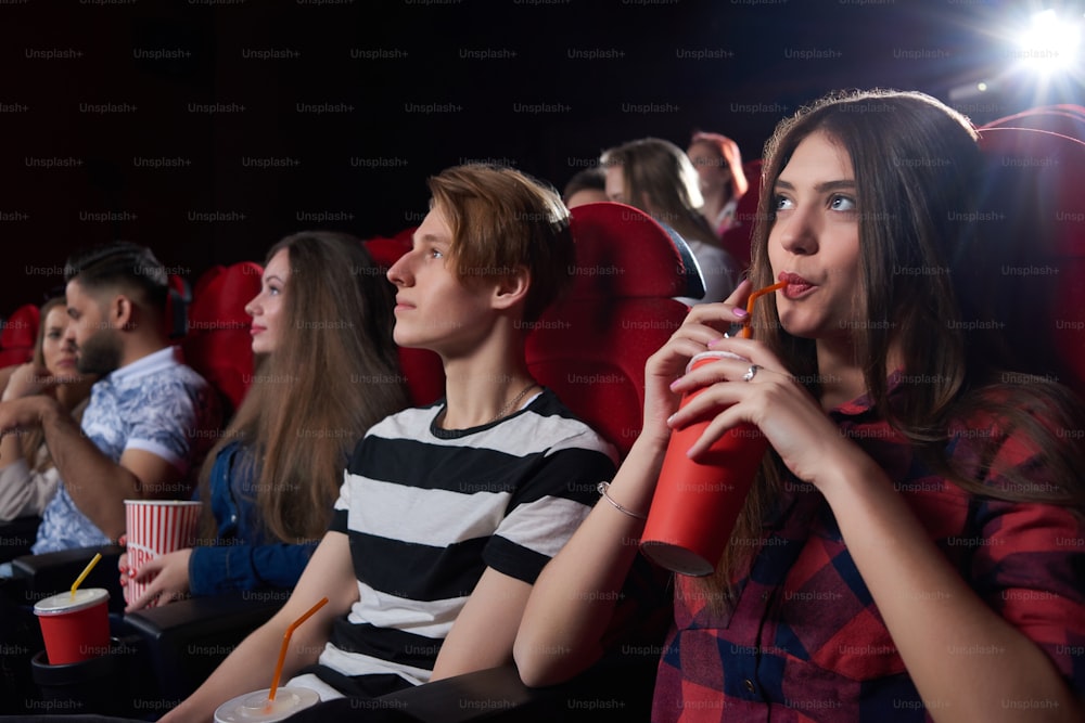 Friends and couples, watching interesting film at big cinema hall with red chairs, serious looking at screen, thinking about movie and drinking cola. Concept of culture and entertainment.