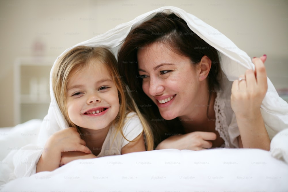 Mãe e filha felizes debaixo do cobertor. Mãe com a filha brincando na cama.