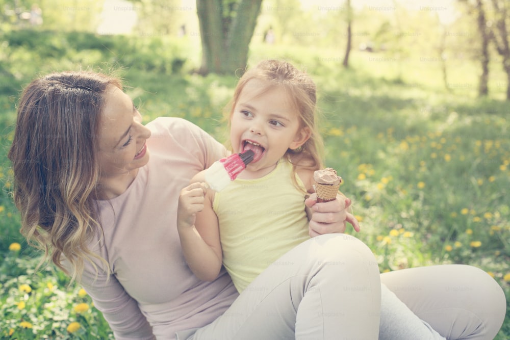 Mother and daughter sitting on the grass and eating ice cream. Happy mother with her daughter in the meadow.