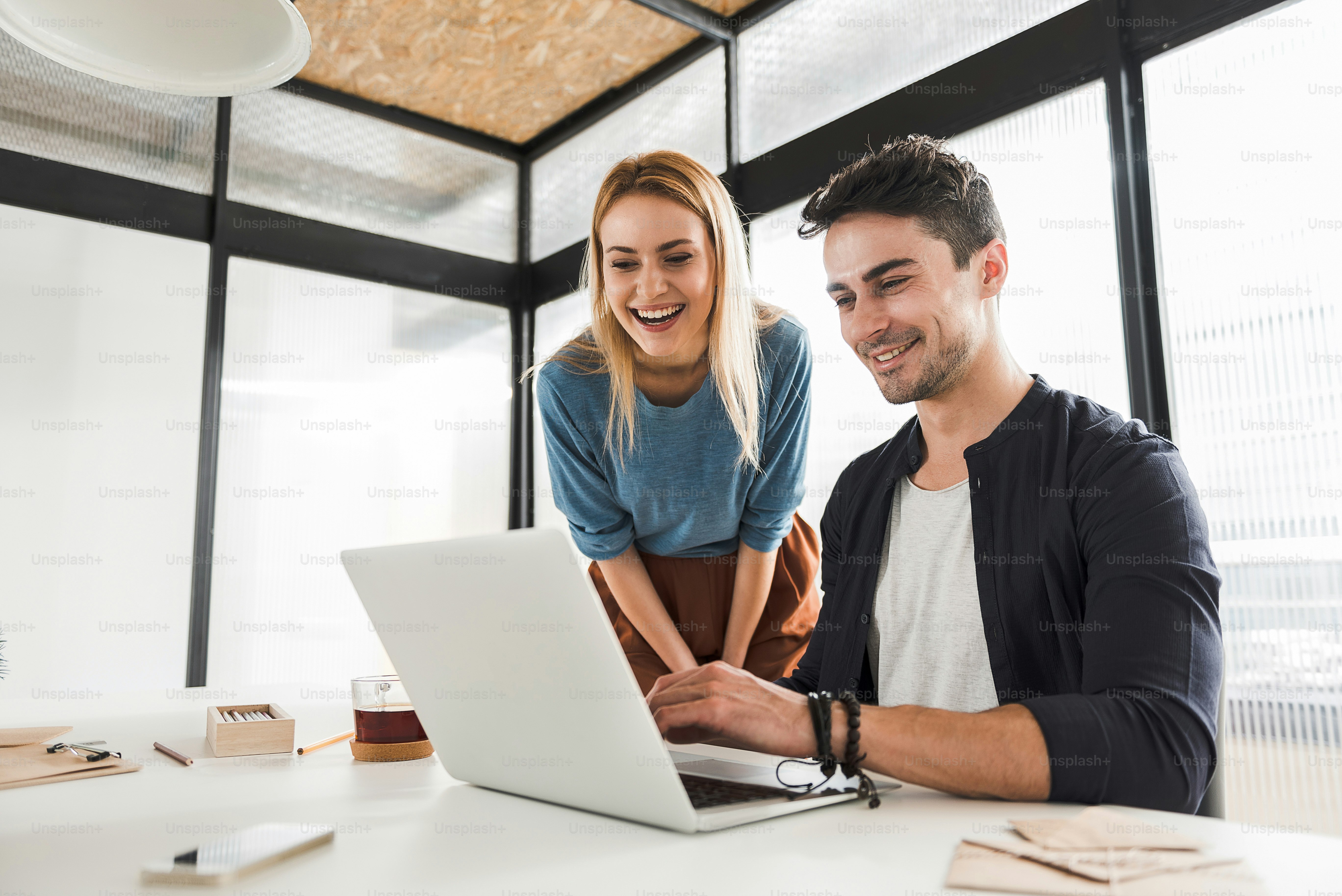 Cheerful man is working on laptop. His female colleague is watching his work with interest. They are looking at screen of device