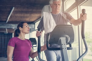 Senior man exercising on stationary bikes in fitness class. Man workout in gym. Senior with personal trainer.