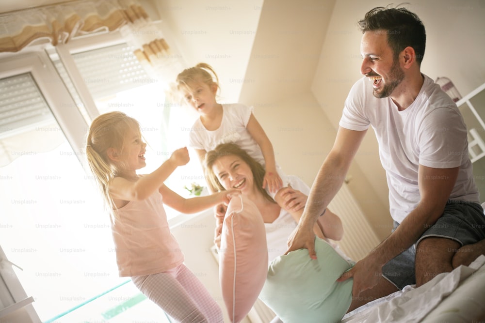 Family having funny pillow fight on bed. Parents spending free time with their daughters. Little girl sitting on mothers shoulders.