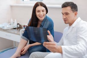 Sie sind gesund. Netter erfreuter angenehmer Arzt, der seiner Patientin ein Röntgenfoto zeigt und erklärt, was darauf ist, während er in ihrer Nähe sitzt