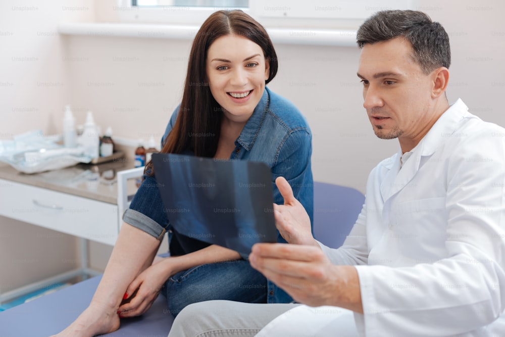 Você é saudável. Nice encantado médico agradável mostrando uma foto de raio X para seu paciente e enquanto explica o que está nela enquanto está sentado perto dela