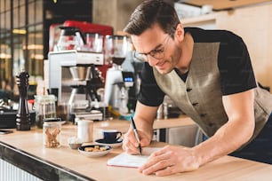 Fröhlicher Barista mit Borsten, der sich Notizen für die Zubereitung köstlicher Getränke in einer gemütlichen Konditorei macht