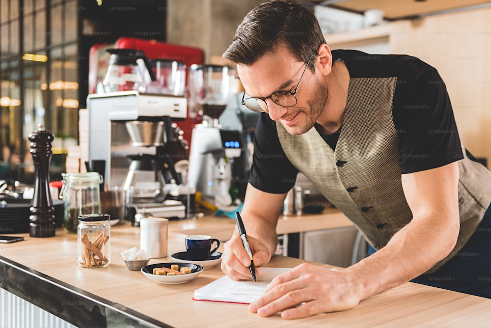 Cheerful bristled barista making notes for preparing delicious beverage in comfortable confectionary shop