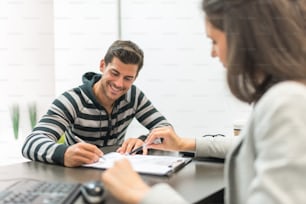 Young cheerful man signing document sitting at desktop with woman in office.