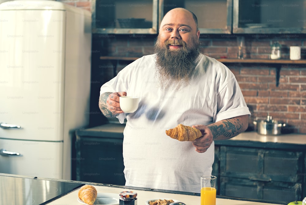Cintura para cima retrato de gordo macho animado bebendo café e comendo croissant em casa. Ele está de pé e sorrindo