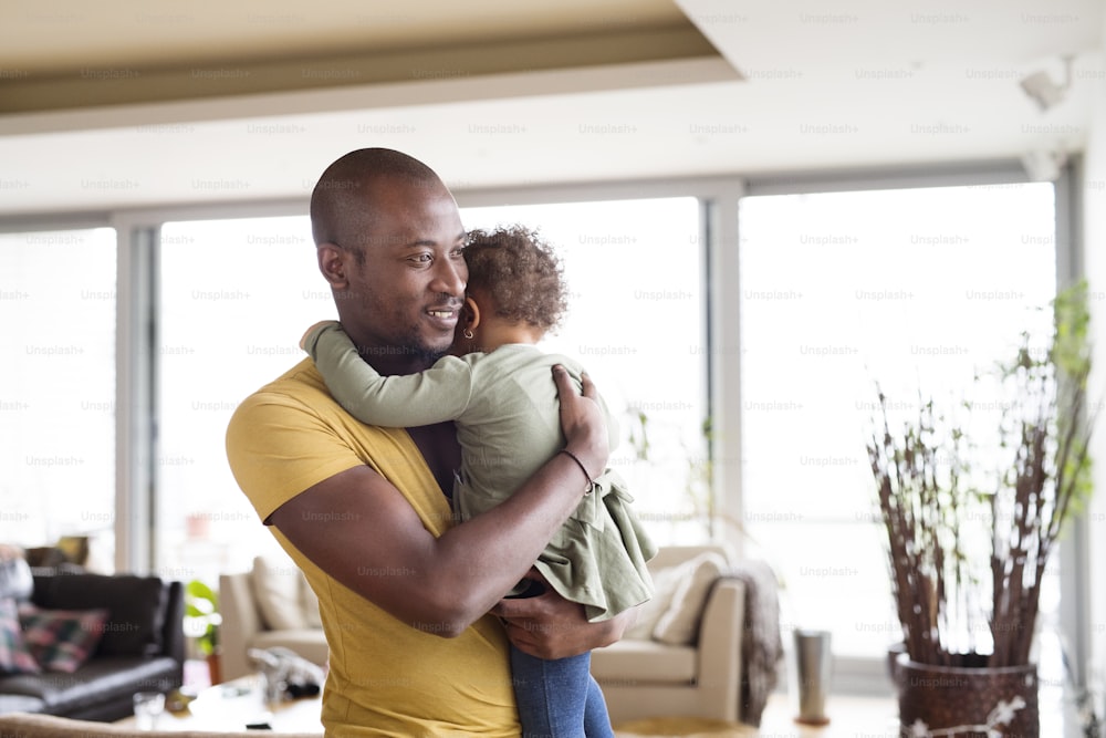 Young afro-american father at home with his cute little daughter holding her in the arms, hugging her.