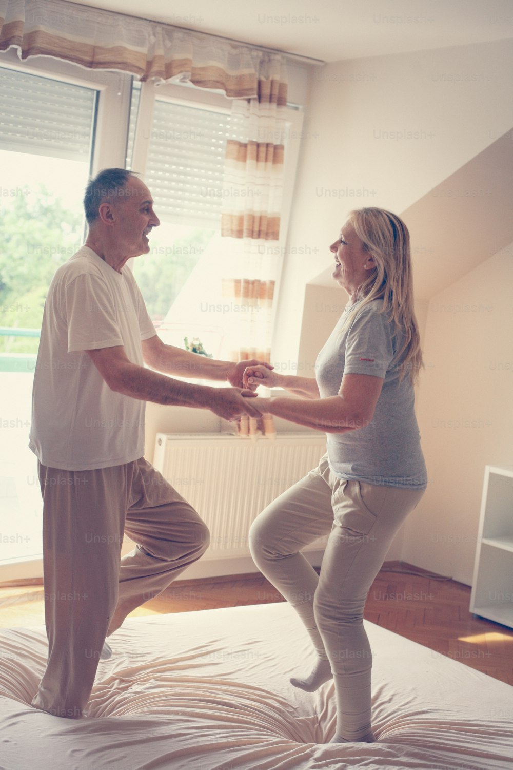 Senior couple dancing and jumping together on bed  holding hands.