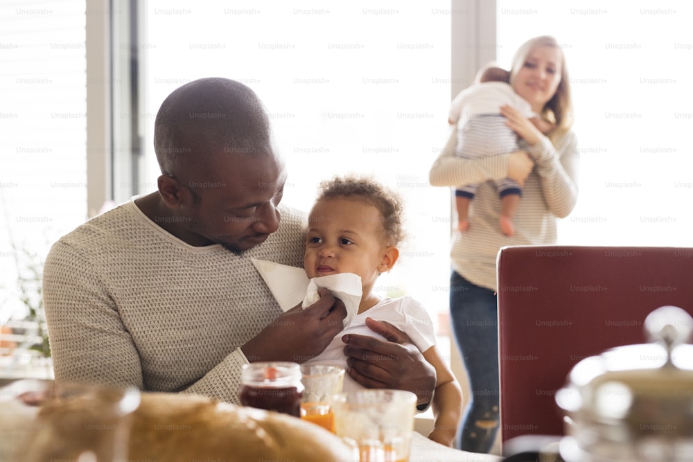 Hermosa familia joven interracial en casa con su linda hija y su pequeño bebé desayunando juntos.
