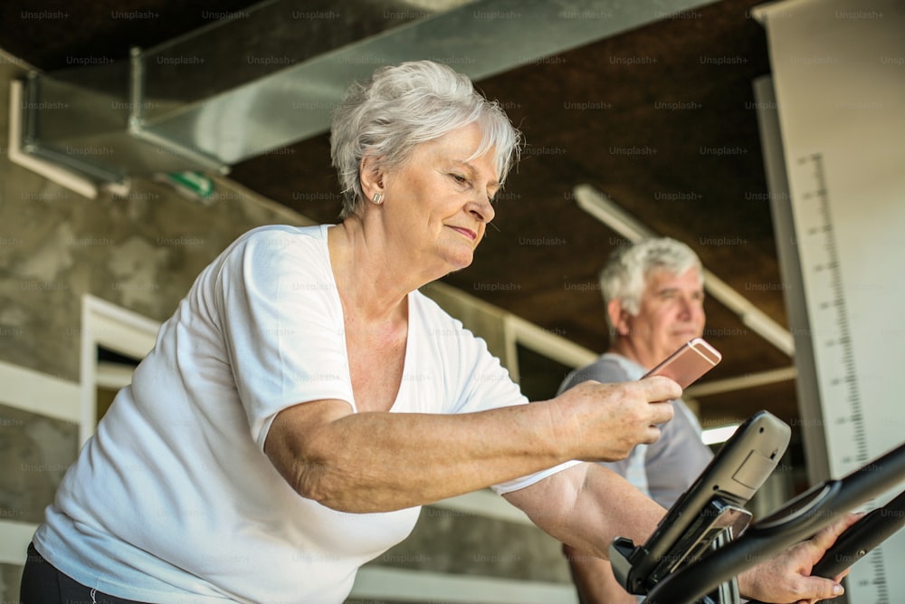 Zwei ältere Leute trainieren auf einem Ellipsentrainer. Ältere Frau mit Smartphone.