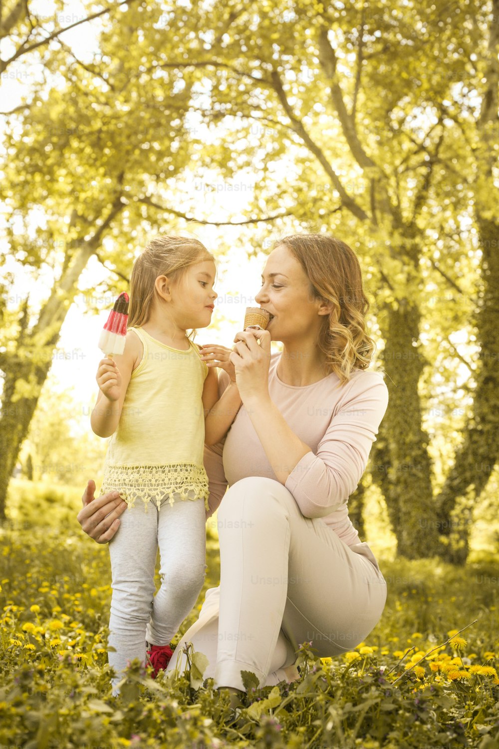Mãe e filha sentadas na grama e comendo sorvete. Mãe feliz e sua filha desfrutando em lindo dia de primavera.