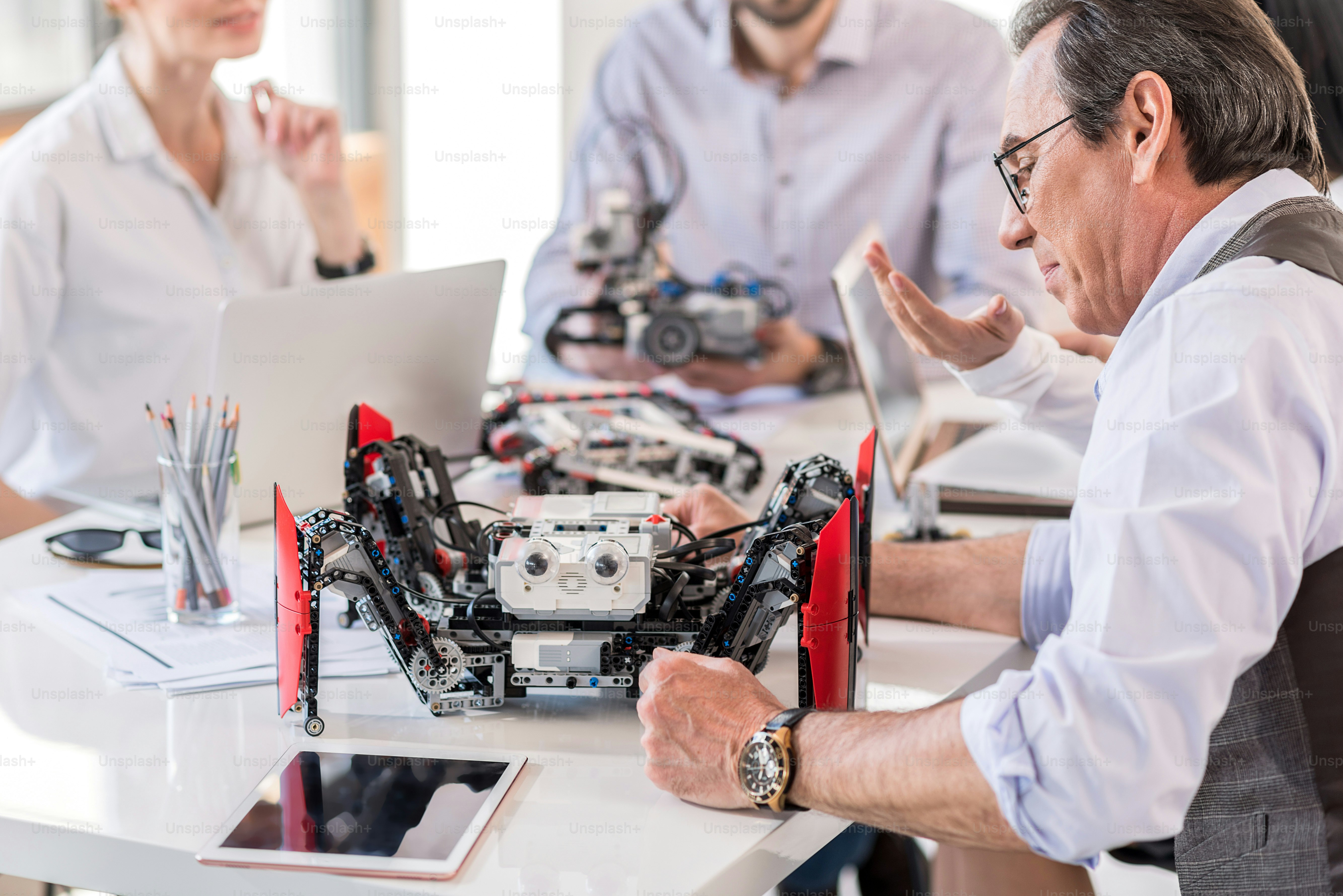Result of work. Selective focus on robot and senior man working with device intently. Young engineers sitting at the table in the background