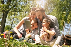 Happy family using digital tablet sitting on the green grass. Family enjoying on the meadow in the spring day.