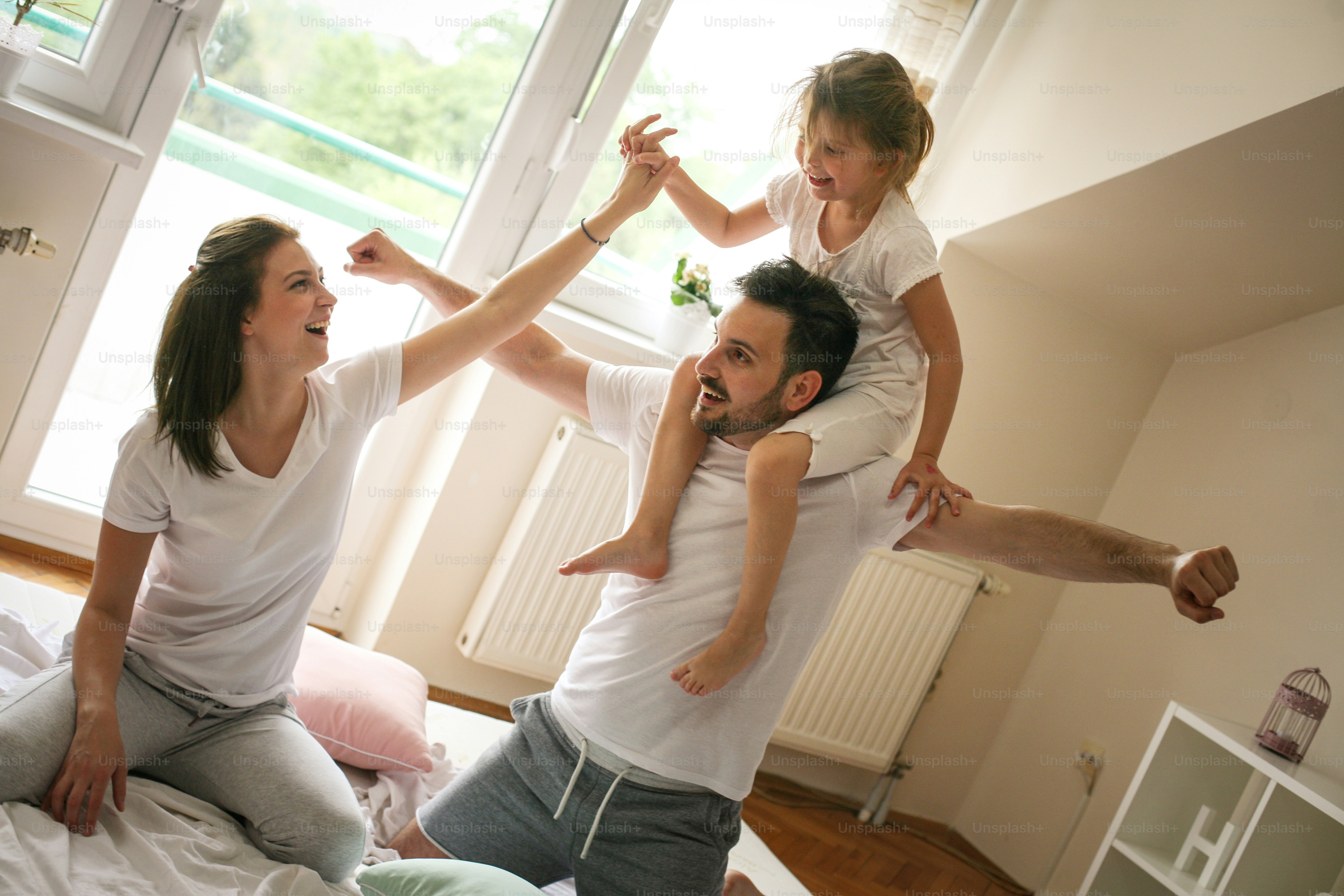Happy family with one daughter spending time at home. Parents playing with their daughter on bed. Little girl sitting on fathers shoulders.