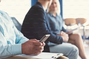 Close up of hands of man sitting on chair in office hall and holding mobile phone by both hands. He is typing sms and leaning arms on clip board lying on laps. Other applicants on background