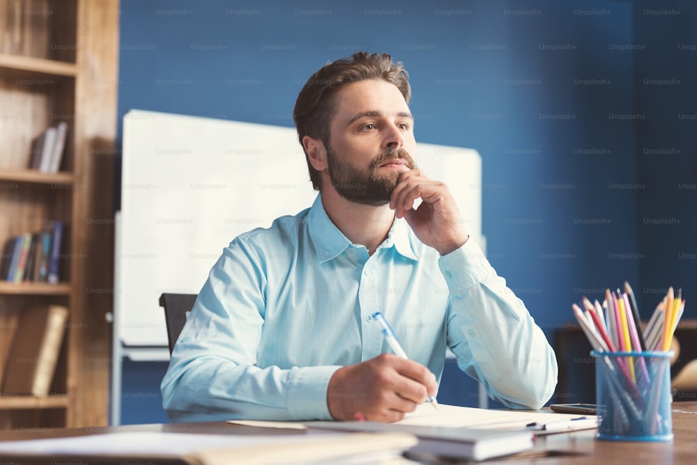 Waist up portrait of pensive young man with beard sitting at table with pen in one hand and thinking over writing creative task. He is leaning on chin by fingers and looking to distance