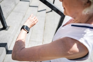 Close up of hand of old serious woman checking pulse. She is standing on big city stairs and looking at tracker on her wrist. Focus on watch